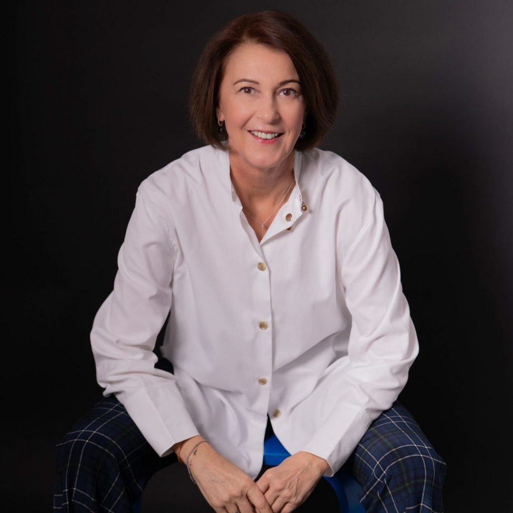 Angelika Schindler-Obenhaus, Geschäftsführerin von ASO Consulting, sitzt lächelnd vor dunklem Hintergrund. Sie trägt ein weißes Hemd und karierte Hose – professionelles, nahbares Porträt für die Website von ASO Consulting mit Fokus auf Leadership, Klarheit und Wirksamkeit.